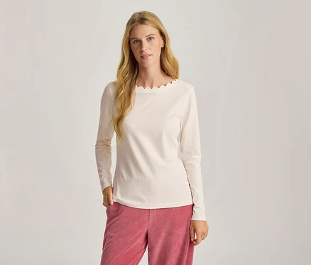 Frau mit langen blonden Haaren trägt ein cremefarbenes Langarmshirt mit Wellenkante am Ausschnitt und eine rosa Cordhose, eine Hand in der Tasche.