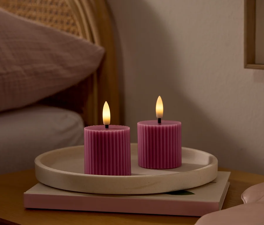 Zwei pinkfarbene, gerippte LED-Kerzen mit leuchtenden Dochten stehen auf einem hellen Tablett auf einem Nachttisch. Daneben liegt eine rosa Schlafmaske. Im Hintergrund ist ein Bett mit einem Flechtkopfteil und einem rosa Kissen zu sehen.