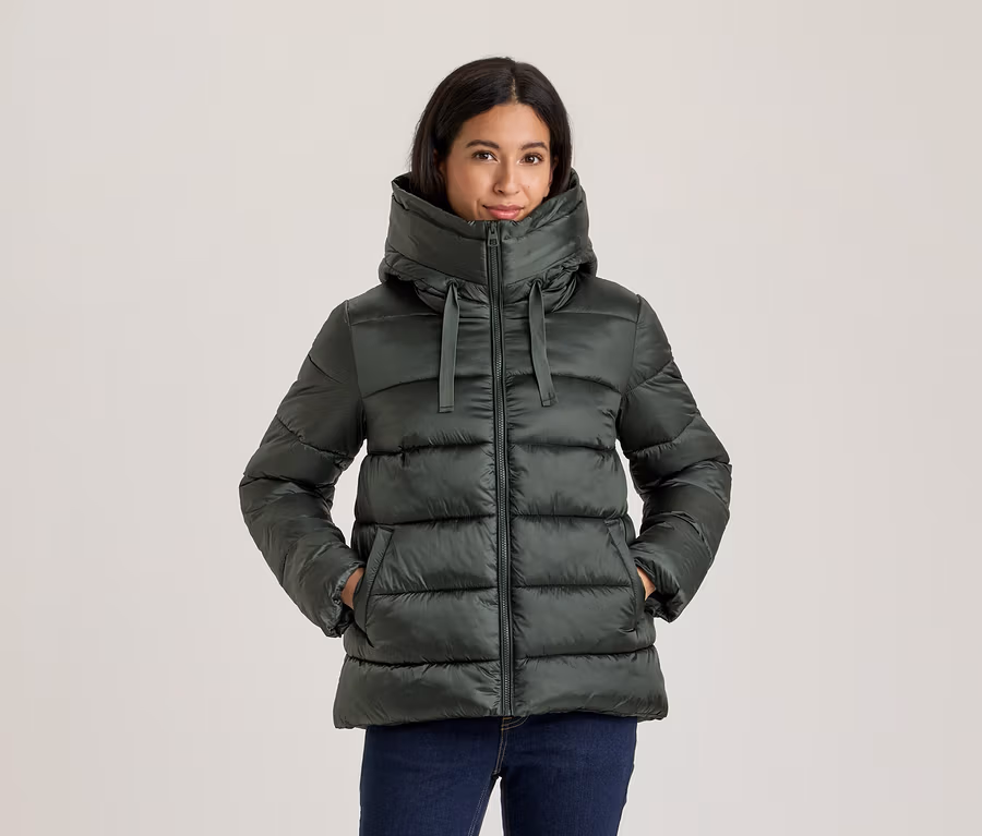Frau in dunkelgrüner Steppjacke mit hohem Kragen und Kapuze, Hände in den Taschen, dazu dunkle Jeans.