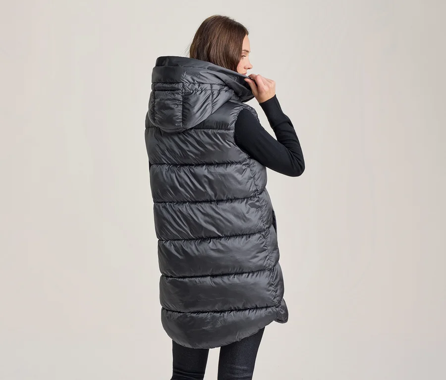 Frau von hinten, trägt eine lange, dunkelgraue, wattierte Kapuzenweste und hält die Kapuze am Hals hoch. Sie trägt ein schwarzes Langarmshirt und dunkle Hosen.