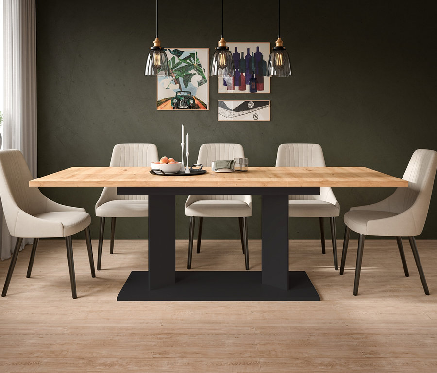 Ein Esszimmer mit einem hellbraunen Holztisch auf schwarzem Sockel, umgeben von sechs beigefarbenen Stühlen. An der dunkelgrünen Wand hängen drei Bilder und drei Pendelleuchten. Auf dem Tisch stehen eine Schale mit Orangen und Kerzen. Links ist ein Fenster mit einem Topf und einer Gießkanne zu sehen.