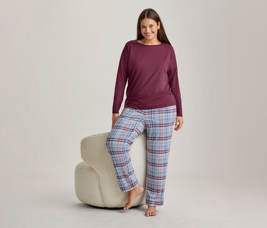 Lächelnde Frau in weinrotem Langarmshirt und hellblau, rot und weiß karierter Pyjamahose, die mit einem Fuß auf einem flauschigen weißen Hocker steht.