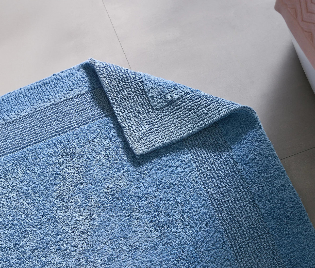 Ein blauer Badteppich liegt auf einem grauen Fliesenboden, eine Ecke ist umgeschlagen. Im Hintergrund ist ein rosafarbenes Objekt zu sehen.