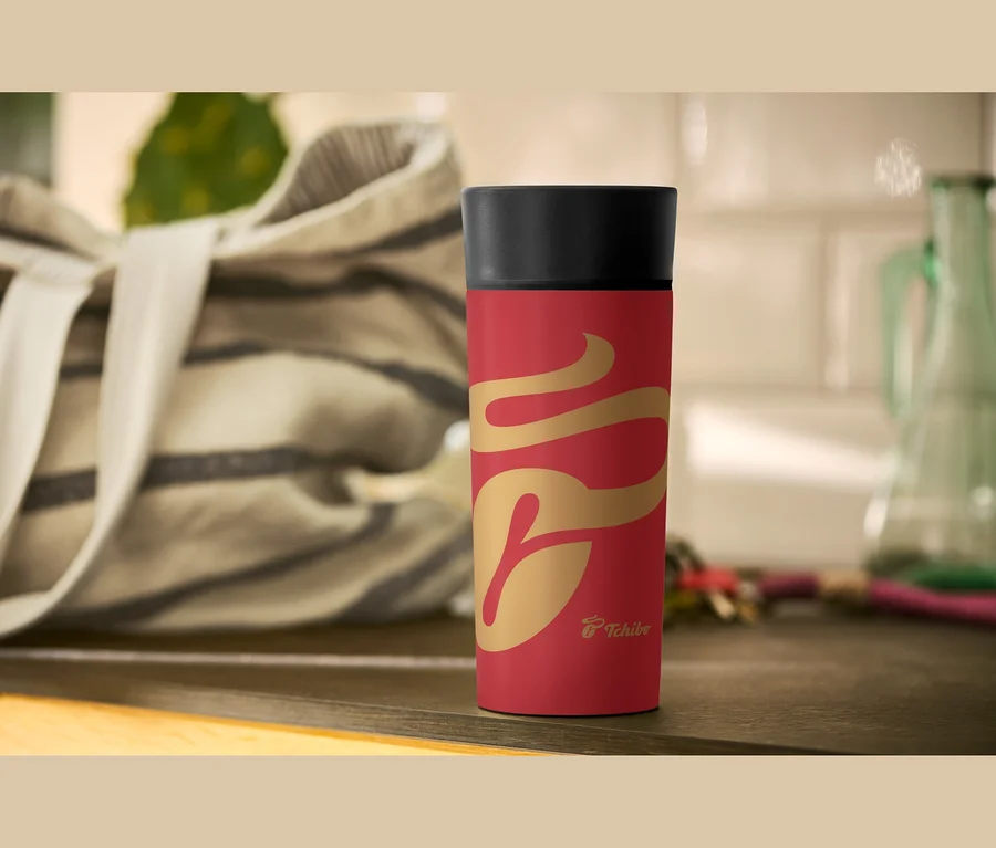 Rote Thermoskanne mit goldenem Muster und schwarzem Deckel auf einer dunklen Holzoberfläche. Im Hintergrund sind eine gestreifte Tasche und ein grüner Glaskrug zu sehen.