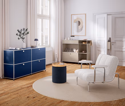 Modernes Wohnzimmer mit Holzboden und weißen Wänden. Links steht ein dunkelblaues Sideboard unter einem Fenster, darauf eine Vase und eine Lampe. In der Mitte liegt ein runder Teppich mit einem zylindrischen Beistelltisch und einer Wasserflasche. Rechts steht ein heller Sessel und ein beiges Rollregal mit Geschirr. An der Wand hängt ein abstraktes Bild.