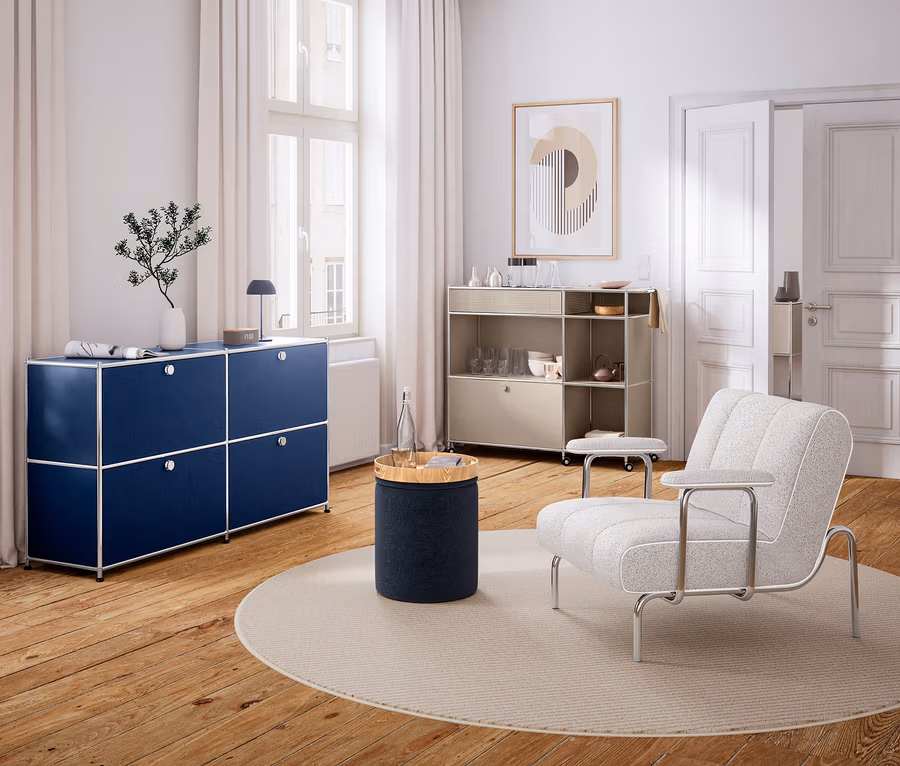 Modernes Wohnzimmer mit Holzboden und weißen Wänden. Links steht ein dunkelblaues Sideboard unter einem Fenster, darauf eine Vase und eine Lampe. In der Mitte liegt ein runder Teppich mit einem zylindrischen Beistelltisch und einer Wasserflasche. Rechts steht ein heller Sessel und ein beiges Rollregal mit Geschirr. An der Wand hängt ein abstraktes Bild.