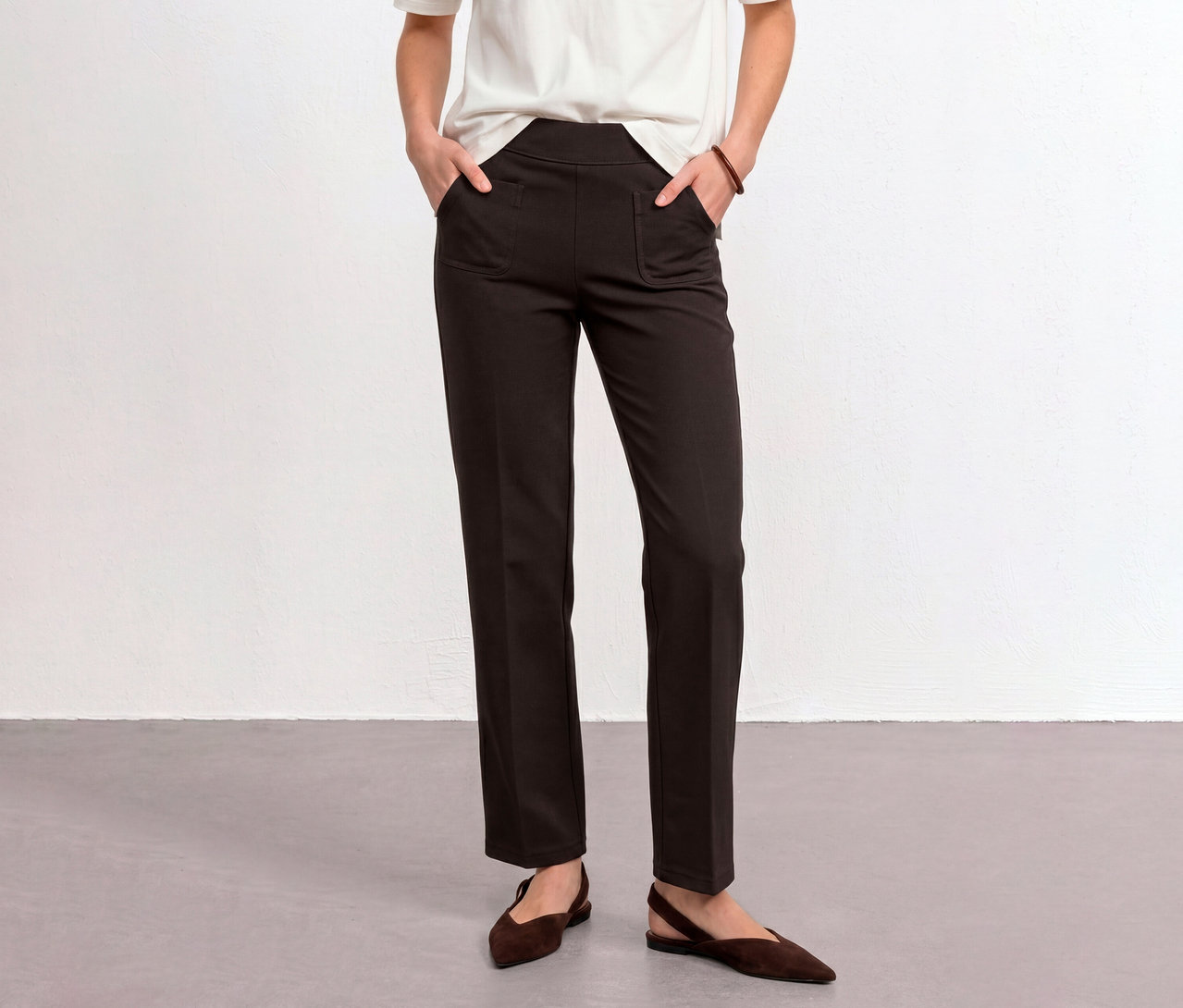 Eine Person trägt eine braune Street One Stoffhose, die Hände in den Taschen, ein weißes T-Shirt und braune flache Schuhe.