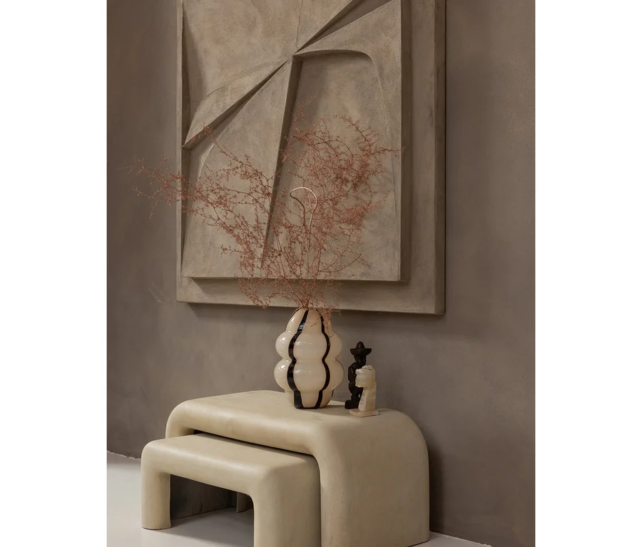 Ein minimalistischer Raum mit einem großen abstrakten Kunstwerk an einer braunen Wand. Darunter stehen zwei ineinander geschobene, hellfarbene, geschwungene Bänke. Auf der oberen Bank steht eine cremefarbene, gewellte Vase mit schwarzen Streifen, die getrocknete Äste enthält, daneben eine kleine schwarz-weiße Figur.