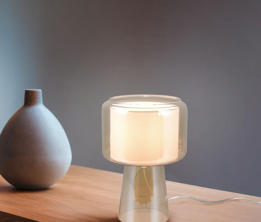 Leuchtende Tischlampe mit klarem Glasfuß und mattem Glasschirm auf einer Holzoberfläche neben einer grauen, birnenförmigen Vase.