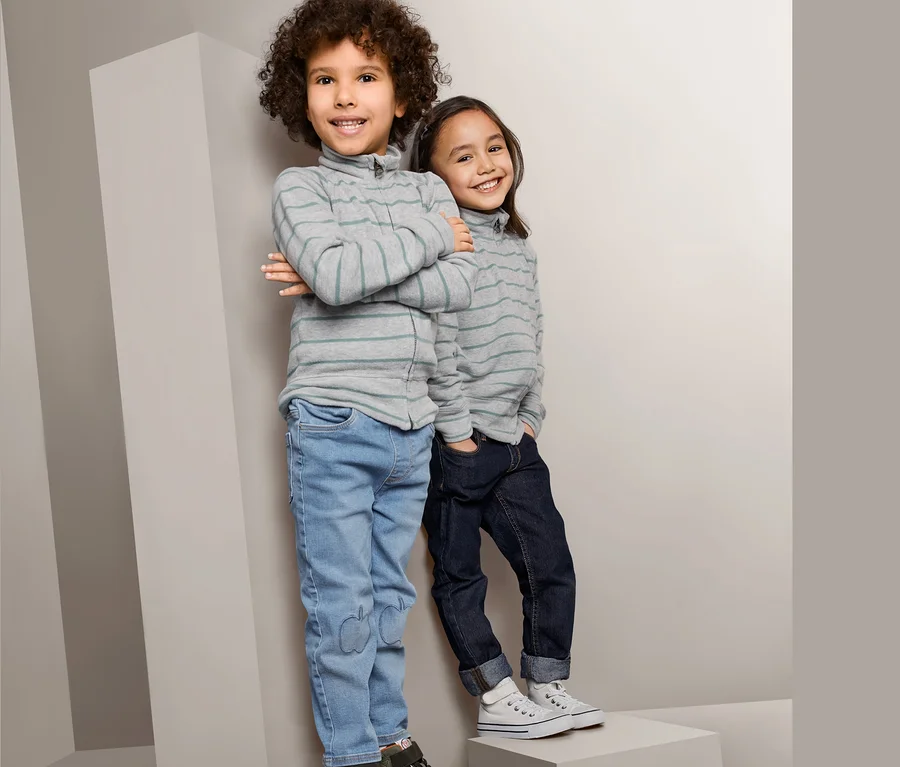 Zwei Kinder stehen auf grauen Blöcken. Der Junge links trägt eine sportive Strickjacke, Jeans und grüne Schuhe. Das Mädchen rechts trägt eine sportive Strickjacke, dunkle Jeans und weiße Schuhe.