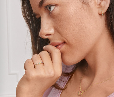 Profil einer brünetten Frau, die Gold-Creolen mit farbigen Zirkonia, eine Gold-Kette und einen Gold-Ring mit farbigen Zirkonia trägt.