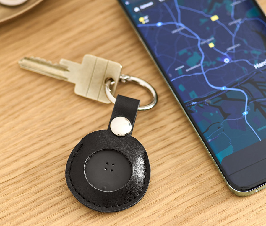 Auf einem Tisch liegen Schlüssel mit einem schwarzen Smart-Tracker mit Anhänger und ein Handy mit einer Karten-App.