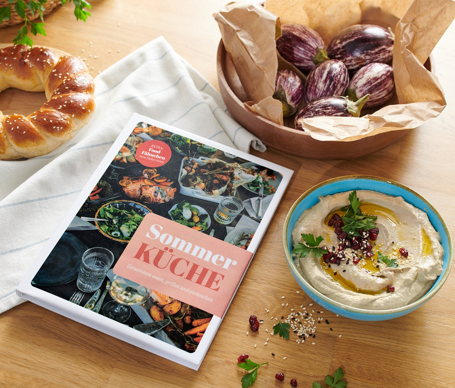 Auf einem Holztisch liegen das Buch »Sommerküche«, ein Kranzkuchen, eine Schale mit Auberginen und eine Schale mit Hummus.