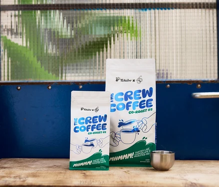 Zwei weiße Kaffeepackungen, eine kleine (250g) und eine große (1000g), mit blau-grünem „Tchibo x 6 THE CREW CO-ROAST #5“-Branding, stehen auf einer Holzoberfläche neben einem kleinen silbernen Becher. Im Hintergrund ist eine blaue Tür mit einem strukturierten Glasfenster zu sehen.