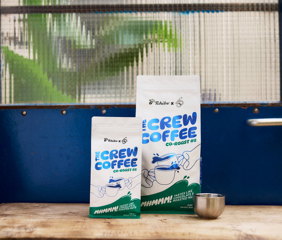 Zwei weiße Kaffeepackungen, eine kleine (250g) und eine große (1000g), mit blau-grünem „Tchibo x 6 THE CREW CO-ROAST #5“-Branding, stehen auf einer Holzoberfläche neben einem kleinen silbernen Becher. Im Hintergrund ist eine blaue Tür mit einem strukturierten Glasfenster zu sehen.
