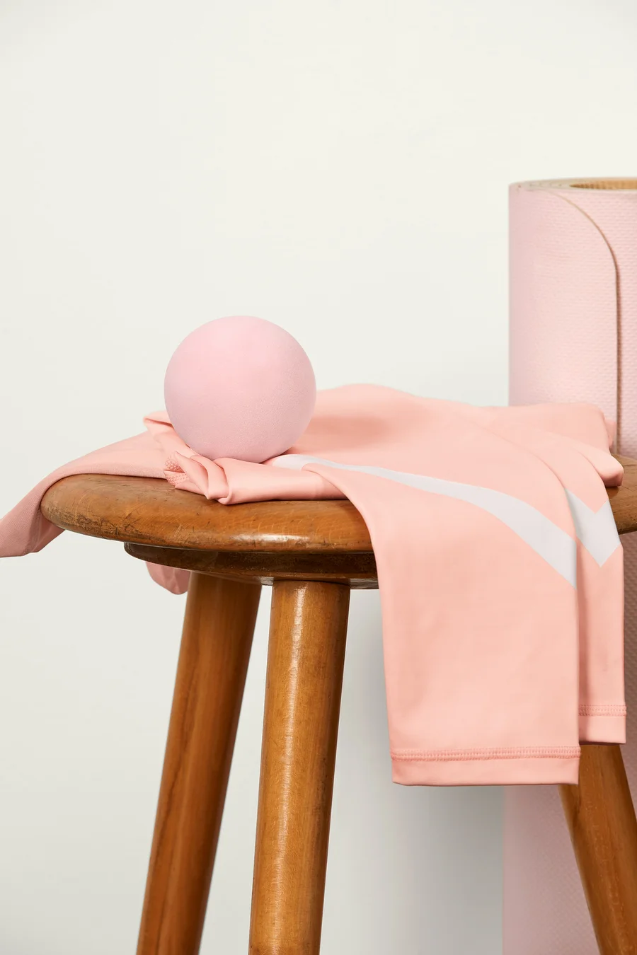 Ein pinker Faszienball liegt auf einer pinken Leggings mit weißem Streifen, die auf einem Holzhocker liegt. Im Hintergrund befindet sich eine pinke Yogamatte.
