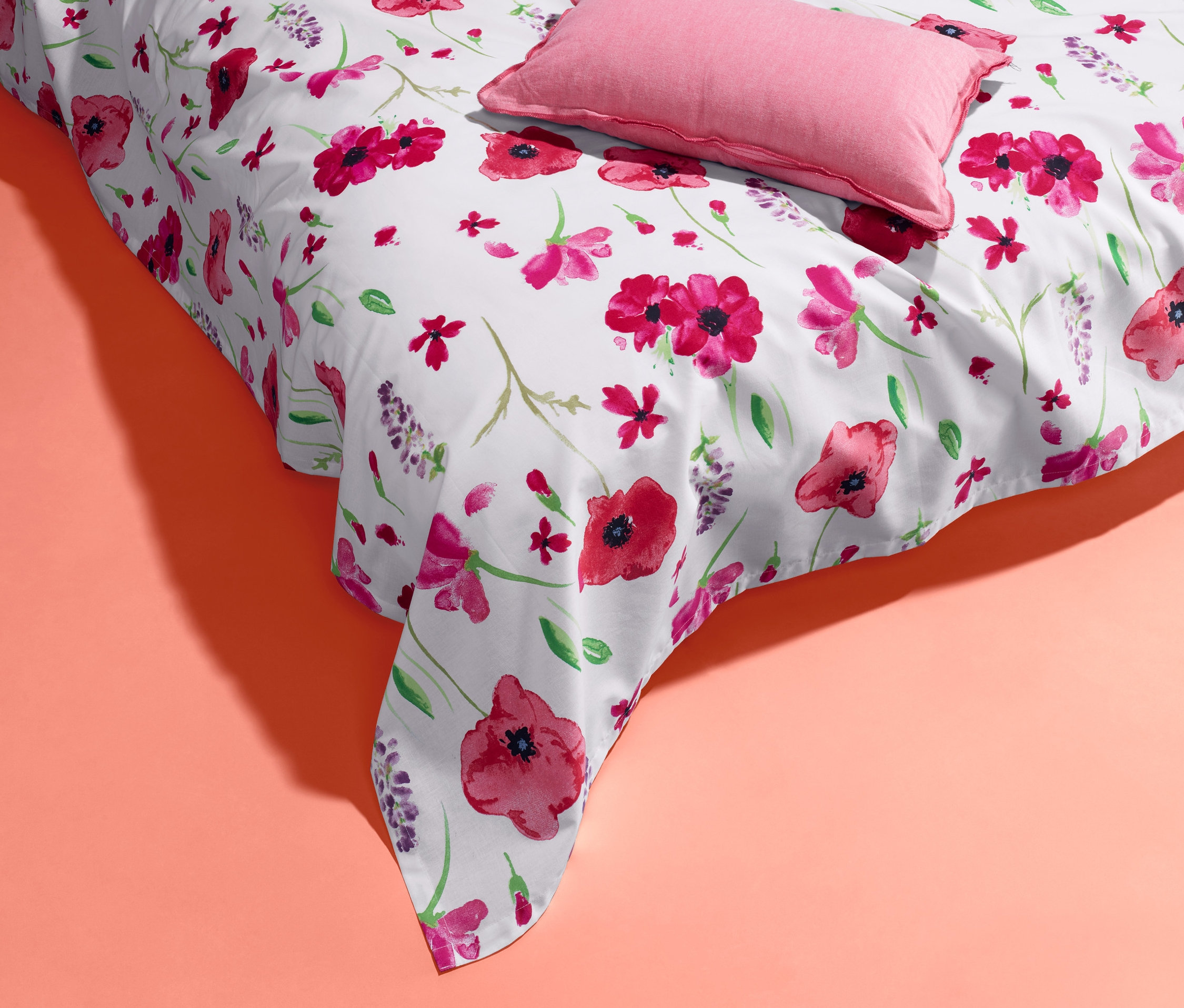 Eine weiße Renforcé-Multifunktions-Überwurf mit Blumenmuster und ein rosa Kissen auf korallefarbenem Hintergrund.