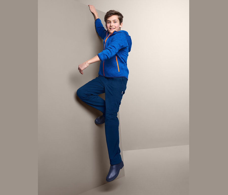 Junge klettert an einer Wand Ecke, trägt eine blaue Softshellhose.