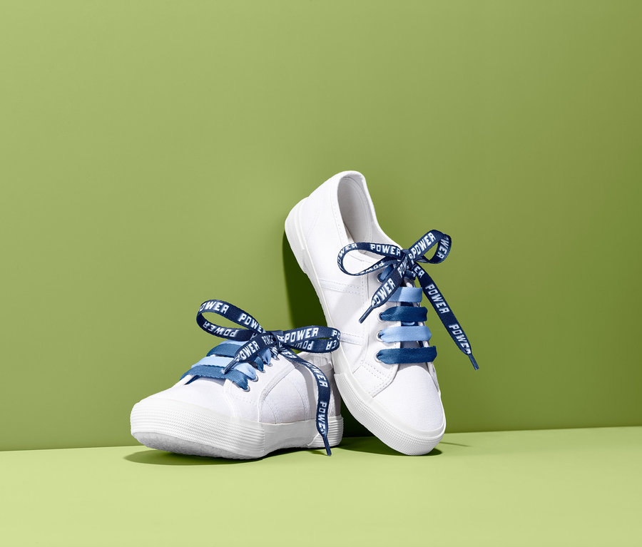 Zwei weiße Schuhe mit blauen Kinder-Schnürsenkel mit Motiv liegen auf einem grünen Hintergrund.