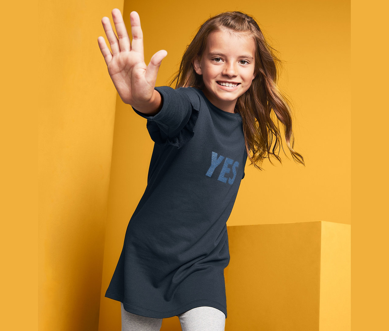 Lächelndes Mädchen winkt und posiert in einem Sweatkleid.