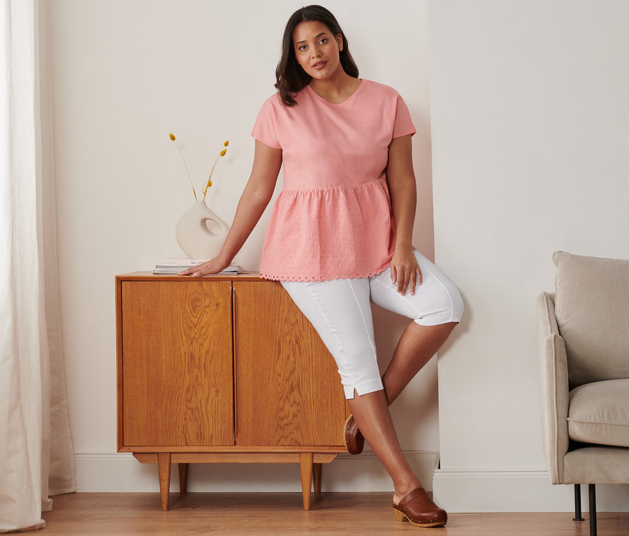 Frau, an Kommode lehnend, trägt korallefarbenes Shirt im Materialmix mit Stickerei, weiße 3/4-Stretchhose und Leder-Clogs.