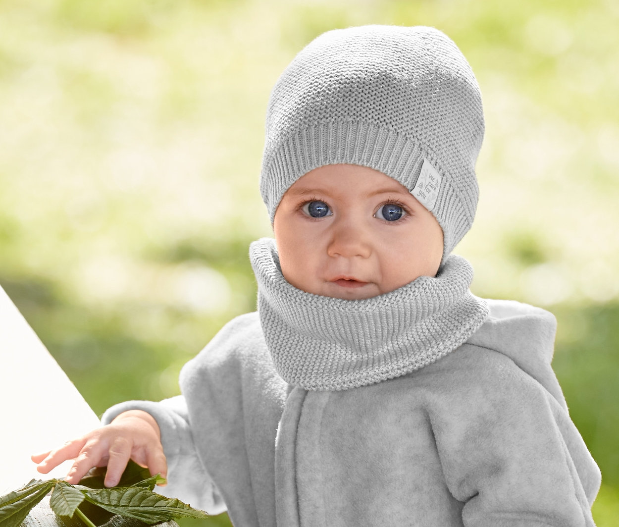 Ein Baby steht an einem Tisch und trägt eine Strickmütze und einen Strickschal, eine Fleecejacke mit recyceltem Material und eine Schlupfhose.