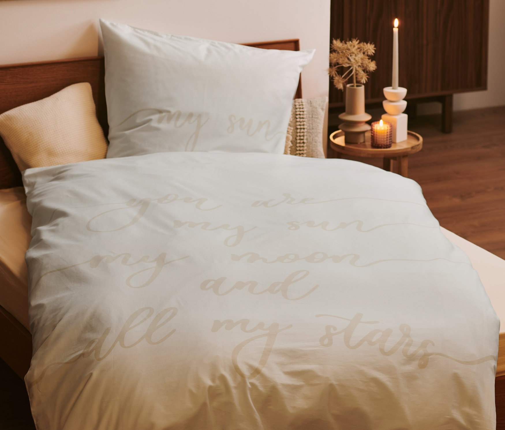 Helle Bettwäsche mit Schriftzug auf einem Holzbett. Im Hintergrund ein Nachttisch mit Kerzen.
