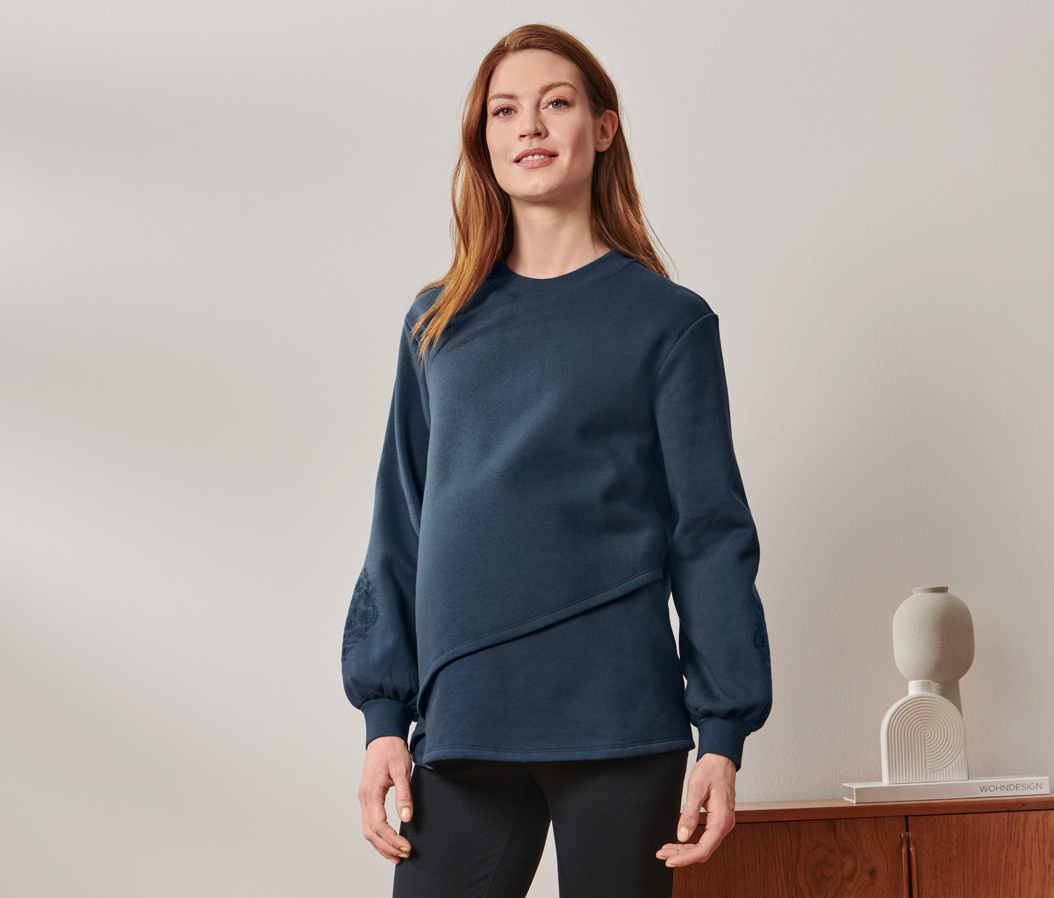 Eine schwangere Frau steht und trägt ein Still-/Umstands-Sweatshirt, schwarze Hosen und Turnschuhe.