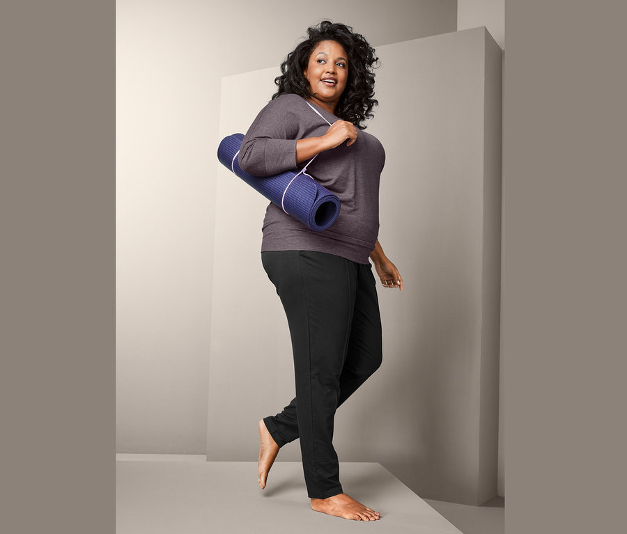 Eine Frau geht eine Stufe hoch. Sie trägt ein 3/4-Sport-und-Yogashirt und eine Sweathose. Über der Schulter trägt sie eine blaue Yogamatte.
