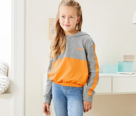 Mädchen steht und trägt einen grau-gelb melierten Cropped-Hoodie, Jeans und Turnschuhe.