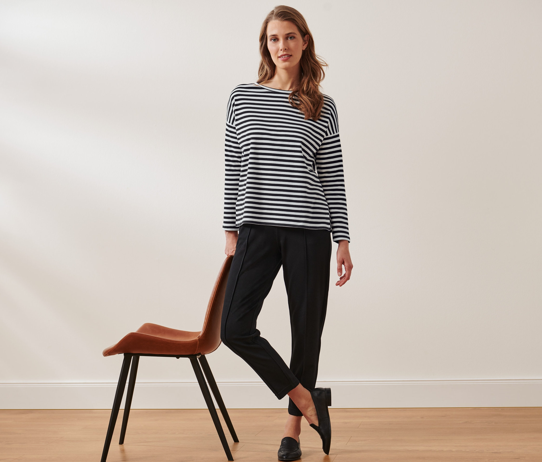 Eine Frau posiert, an einen braunen Stuhl gelehnt, in einer schwarzen Hose und einem gestreiften Langarmshirt.