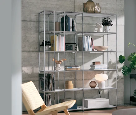 Ein offenes Regal Metall »CN3« in Weiß mit einer Tischleuchte mit Glasschirm und einem Relaxstuhl.
