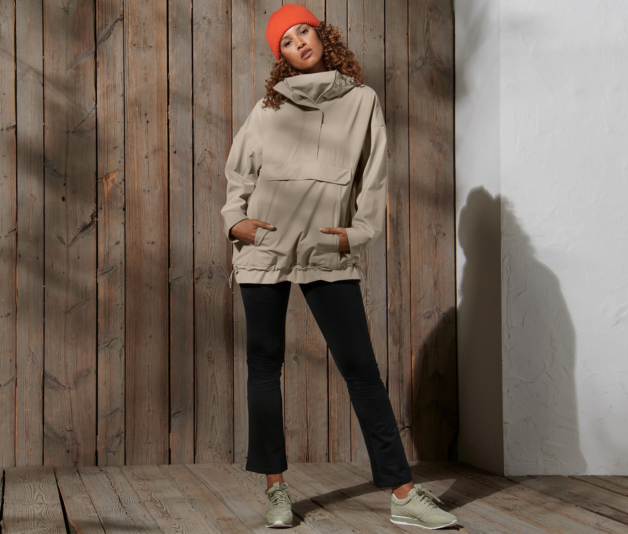 Frau posiert vor einem Holzhintergrund und trägt eine orangefarbene Mütze und eine beige Oversized-Funktionsregenjacke.