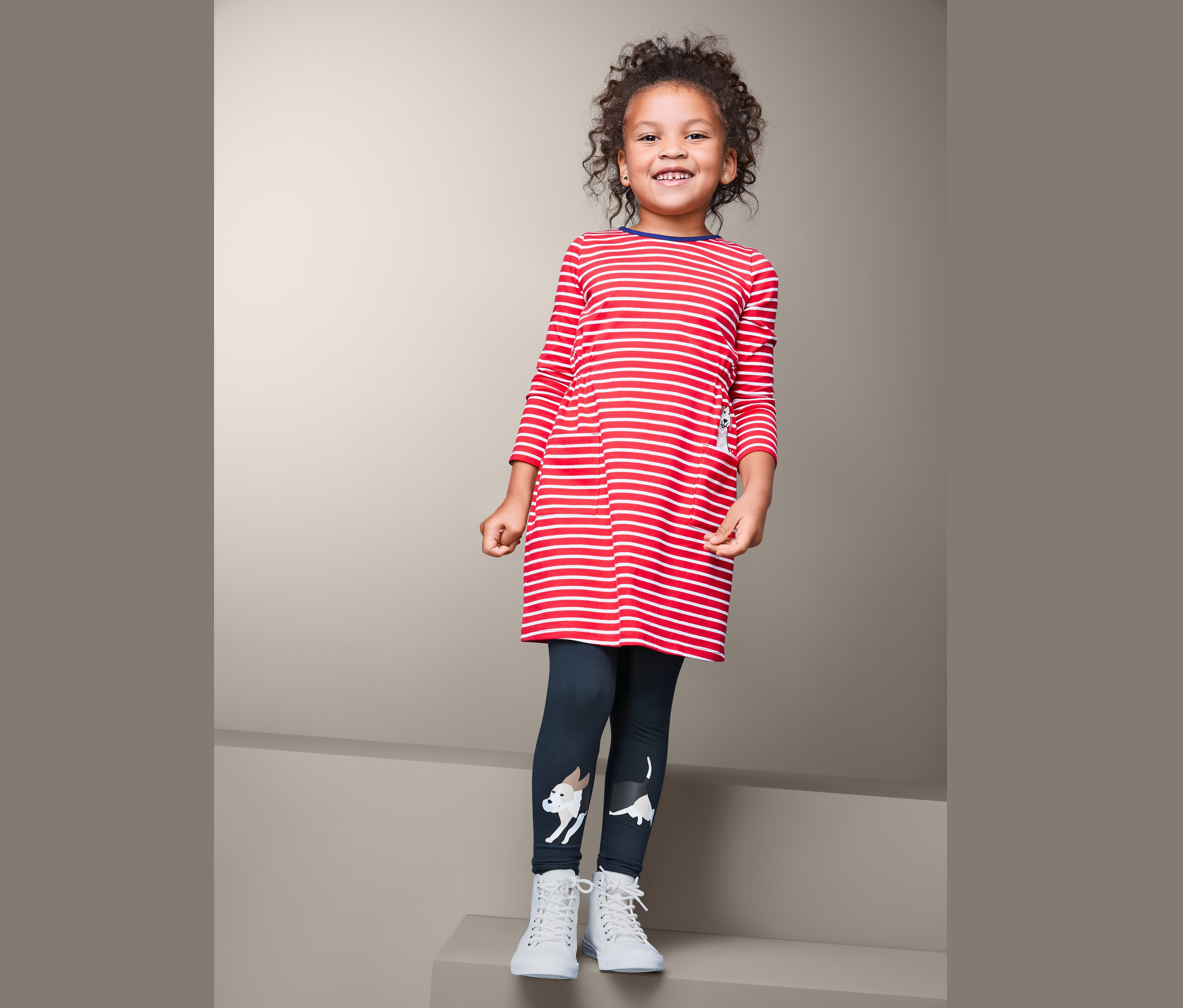 Lächelndes Mädchen posiert in einem rot-weiß gestreiften Langarmkleid und 2 Leggings mit Hundemotiv.