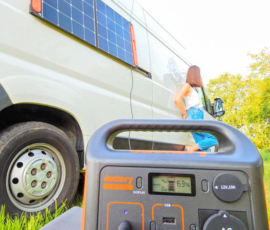 Eine Frau lehnt an einem weißen Lieferwagen mit einem Solarpanel. Im Vordergrund befindet sich ein Jackery Explorer 240.