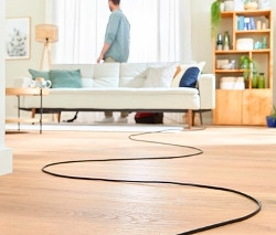Ein Mann steht im Wohnzimmer hinter einem weißen Sofa. Auf dem Holzboden ist das Kabel von einem Bosch Bodenstaubsauger »BGD38RD1H« mit Beutel zu sehen.