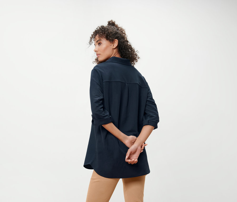 Eine Frau steht mit dem Rücken zur Kamera und hat die Hände hinter dem Rücken verschränkt. Sie trägt eine dunkelblaue Piqué-Jerseybluse und Khaki-Hosen.