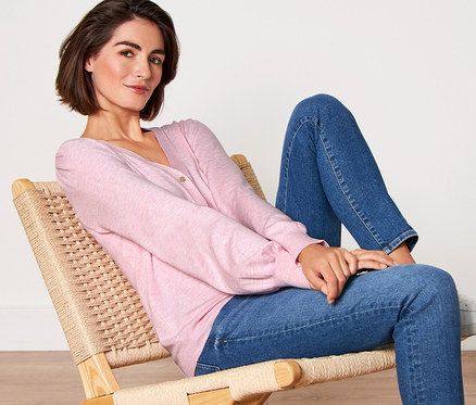 Frau sitzt in einem Stuhl und trägt einen roséfarbenen Feinstrick-Cardigan, Jeans und weiße Turnschuhe.