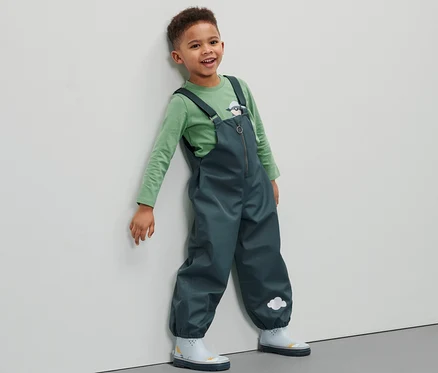 Ein Junge in einer Kleinkind-Regenhose lehnt an einer Wand.