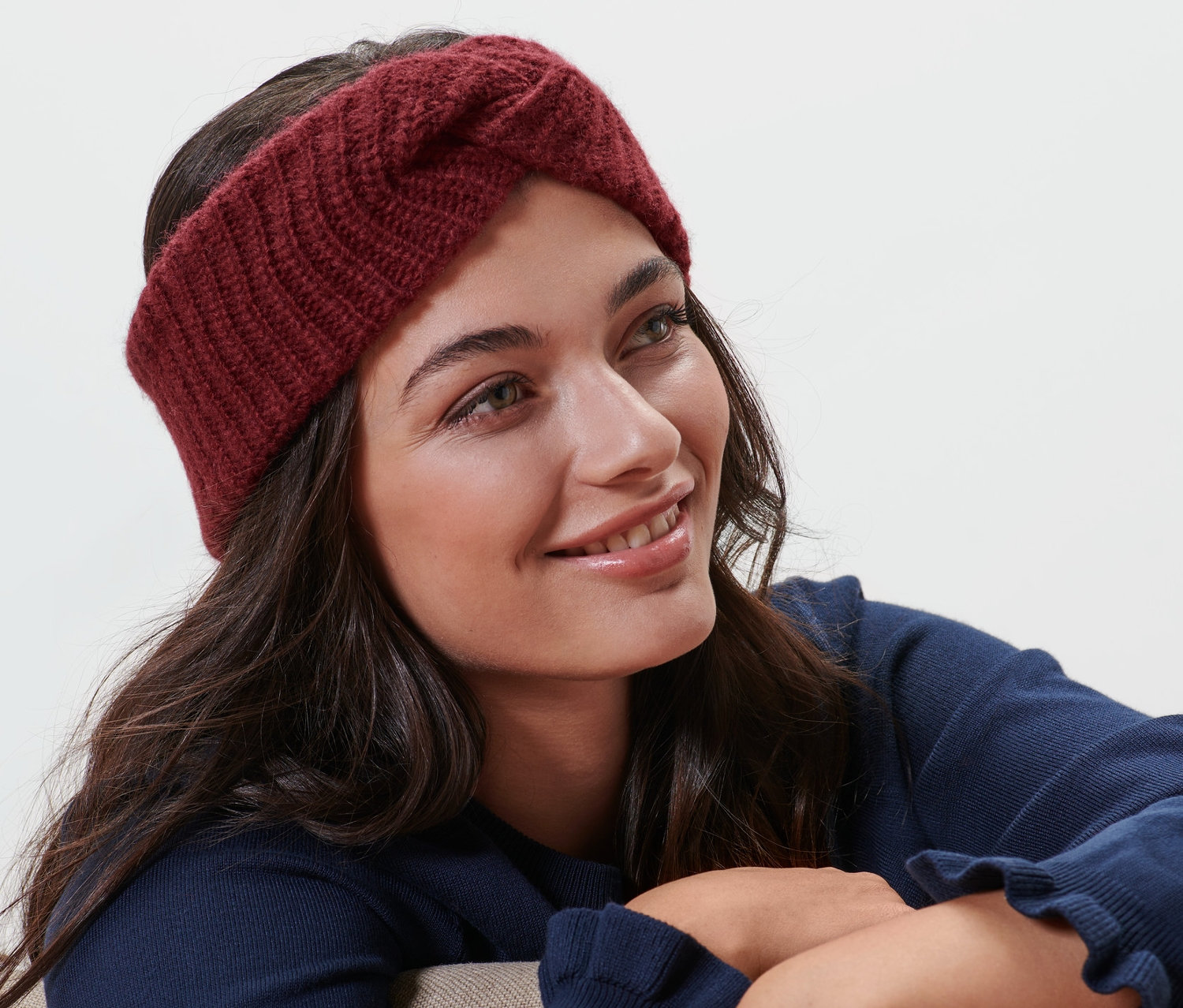 Frau mit braunen Haaren und einem bordeauxroten Stirnband mit Wolle sitzt auf einem beigen Stuhl und lächelt.