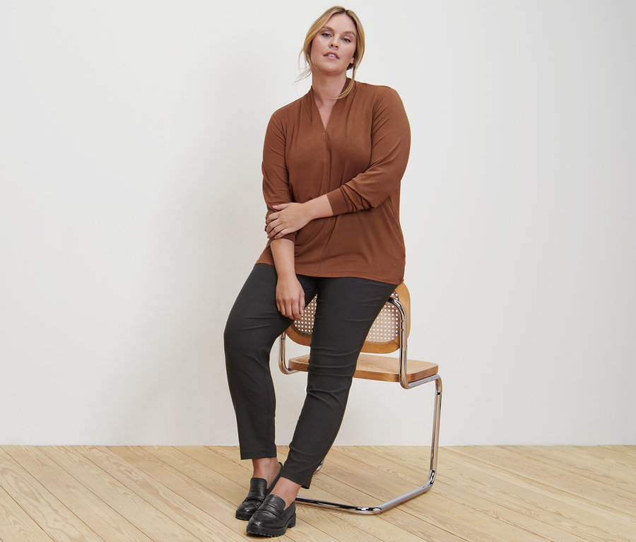 Eine Frau sitzt auf einem Stuhl und trägt ein cognacfarbenes Langarmshirt mit V-Ausschnitt, eine Bengalin-Hose und Leder-Loafer.