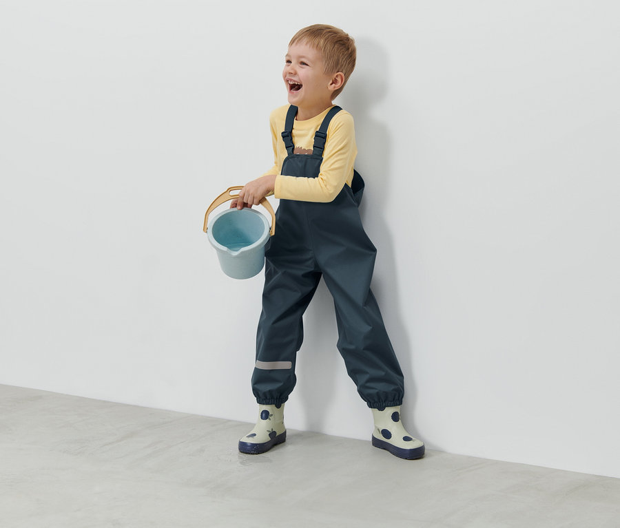 Lachender Junge hält blauen Eimer und trägt dunkelblaue Kleinkind-Regenhose.