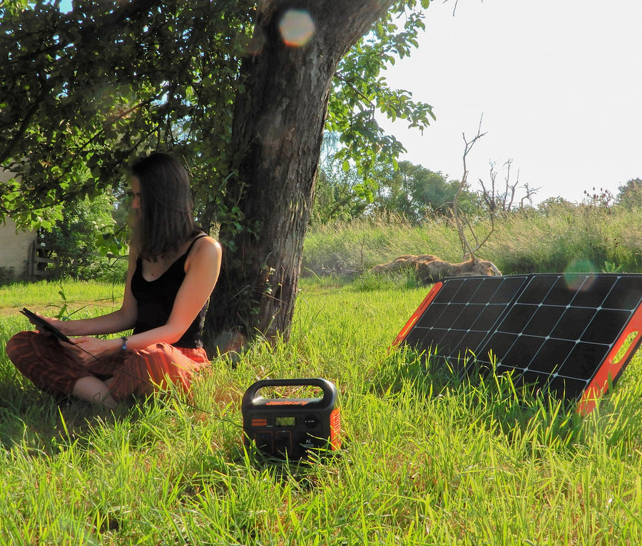 Eine Frau sitzt unter einem Baum und benutzt ein Tablet. Daneben befinden sich ein Solarpanel und ein Jackery Explorer 240.