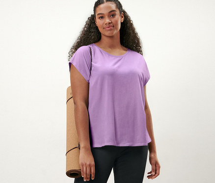 Frau steht auf Holzboden, trägt ein fliederfarbenes Sportshirt und eine Bodyforming-Sporthose, eine Yogamatte unter dem Arm.