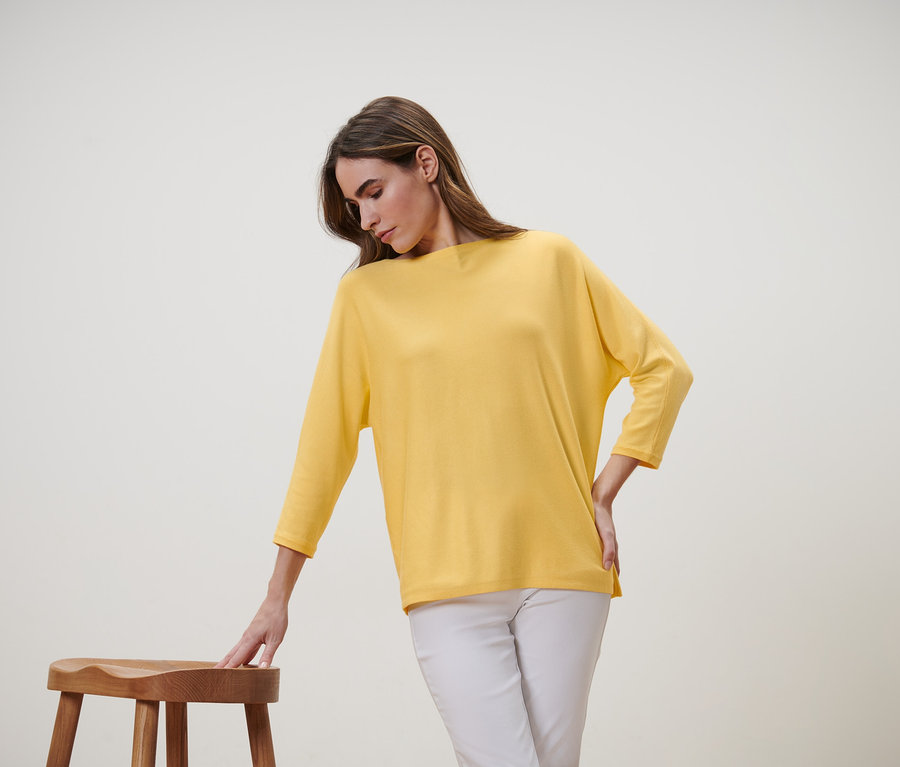 Frau beugt sich zu einem Holzhocker, trägt einen gelben Feinstrickpullover und eine weisse Hose.