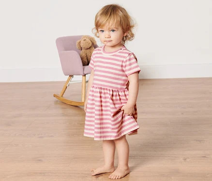 Mädchen steht auf dem Boden und trägt ein Baby-Jerseykleid mit rosa Streifen.