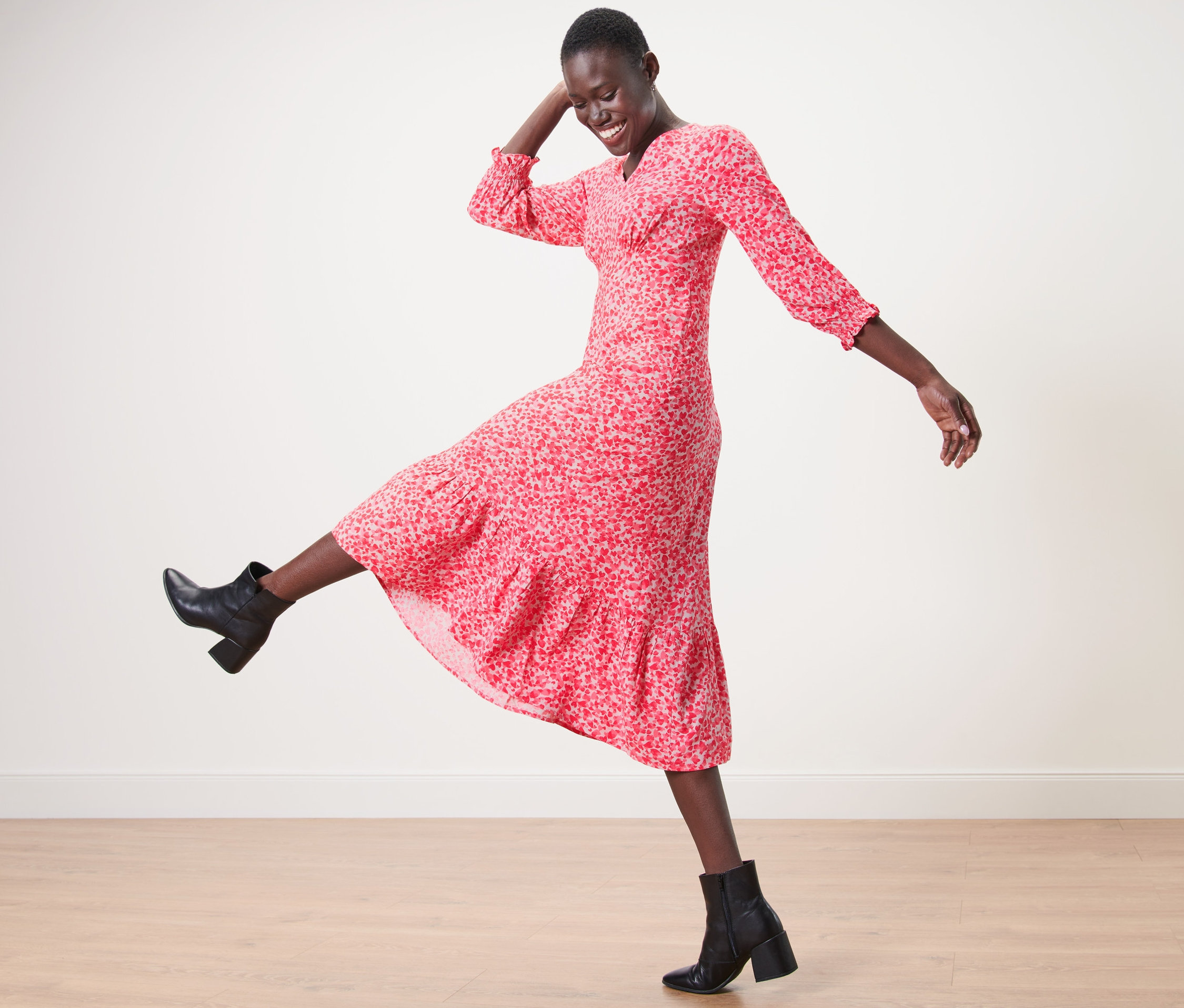 Frau in Maxikleid mit Print und schwarzen Stiefeletten posiert auf Holzboden.