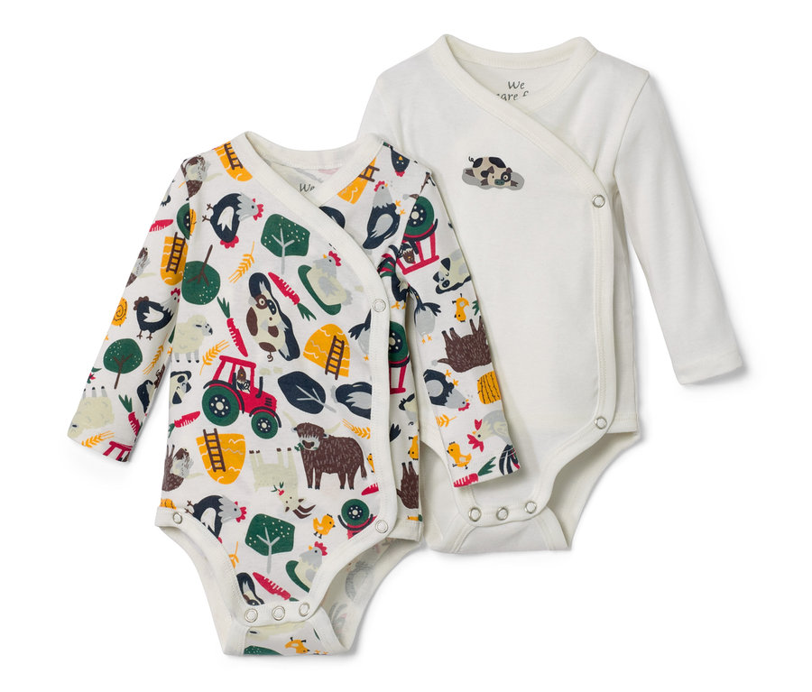 Zwei Baby-Langarmbodys liegen nebeneinander. Einer ist weiß mit Bauernhofmotiven, der andere ist weiß mit einem kleinen Kuhmotiv.