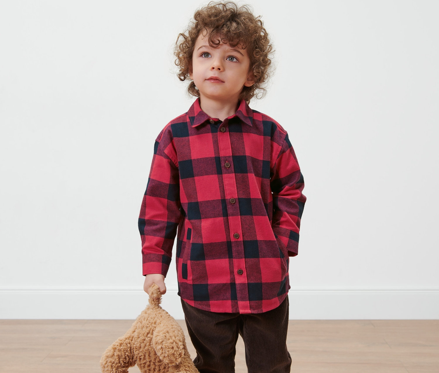 Ein Junge mit lockigen Haaren trägt ein Kinder-Flanellhemd und hält einen Teddybären.