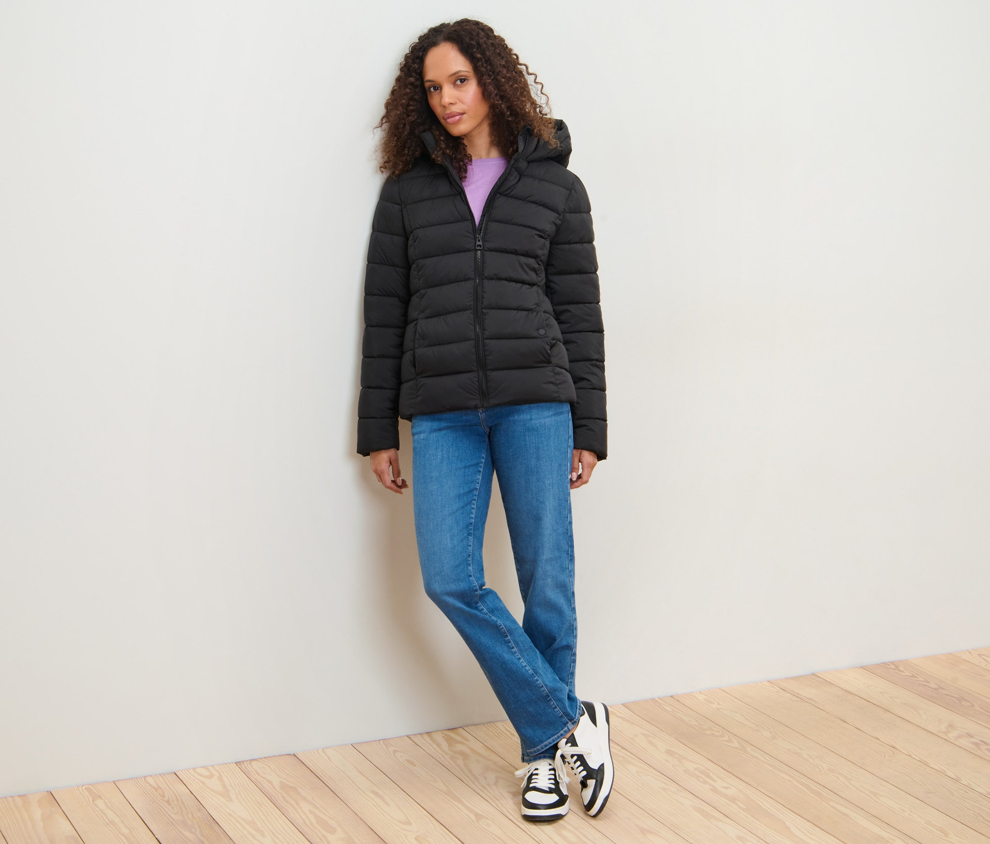 Frau, an eine Wand gelehnt, trägt eine Steppjacke mit Kapuze.